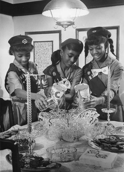 African American junior Girl Scouts pouring fruit juice into a punch bowl as they prepare to entertain their parents.