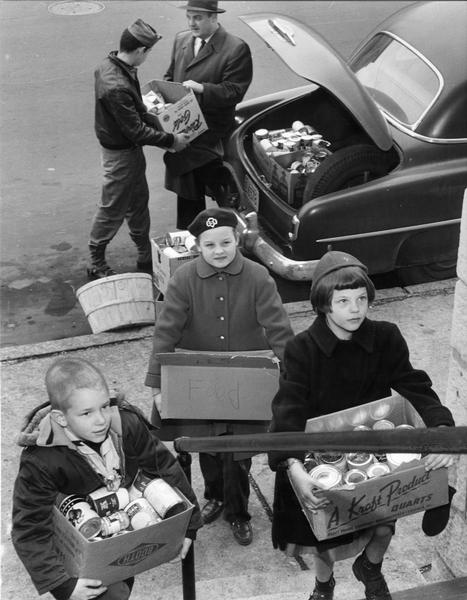 Elevated view of a Cub Scout, Girl Scout, and Brownie Scout standing on the sidewalk bringing boxes of food to St. Vincent de Paul to be distributed to recently arrived Hungarian refugees.