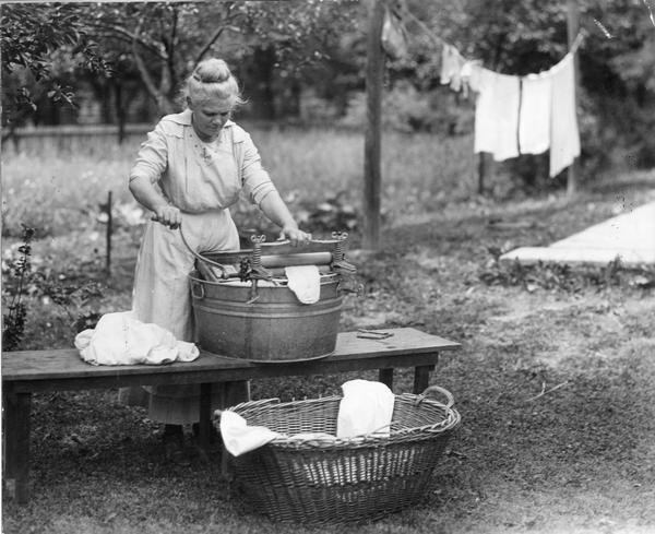 Woman washing clothes outdoors with a washtub, washboard, wringer, clothespins and clothesline.
