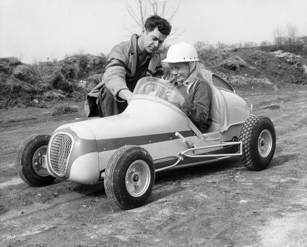 Father demonstrates controls of a mini-race car as his daughter sits in the car.