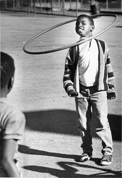 A boy is twirling a "hula-hoop" around his neck while another child is watching from the left foreground.