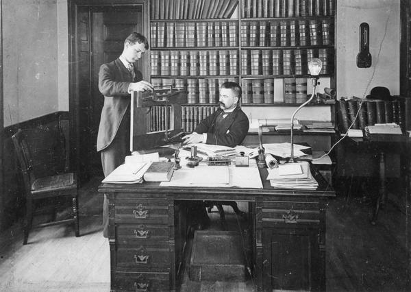 Patent attorney Charles L. Goss (seated), with clerk Arthur Muerless in the law offices of Winkler, Flanders, Smith, Bottum, & Vilas.
