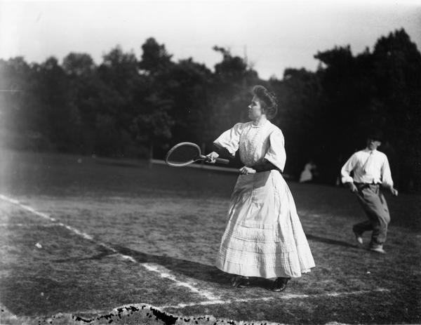 Mixed doubles tennis game outdoors at the beginning of the 20th century, with a woman clad in long dress and upswept hair. A man wearing a tie is running behind her.