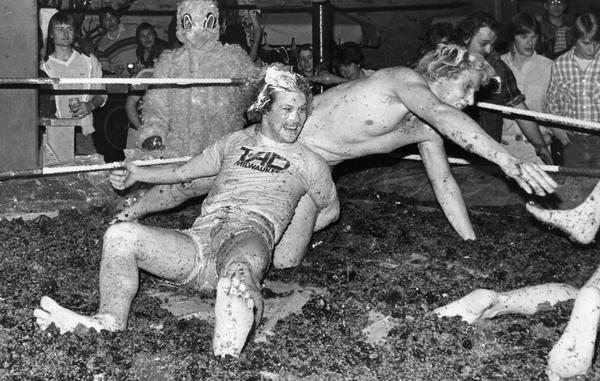 Onlookers watch as participants flail around in a mud pit wrestling ring. One spectator wears a bird costume.