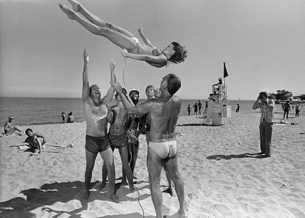 A bikini-clad woman snaps a self-portrait with a long cable release attached to a camera as she is tossed into the air by a group of men on the beach. Other people on the beach are watching and taking snapshots, and there is a lifeguard on a lifeguard station in the background.
