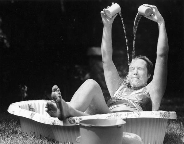 Woman stays cool by sitting in a small plastic pool while pouring cups of water on her head.