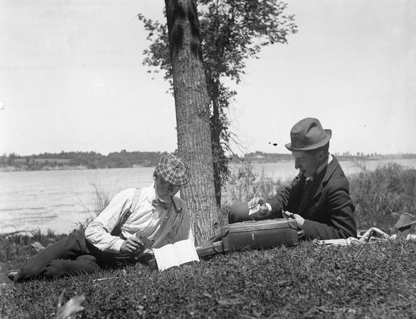 Two men smoking pipes and playing cards in the grass along a riverbank.