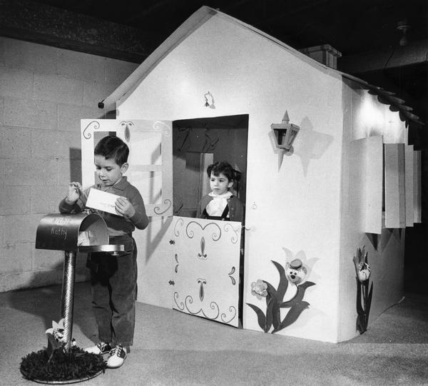 A young girl watches out the door of her playhouse while a young boy retrieves letters from the play mailbox.