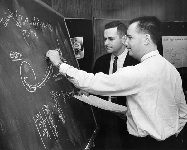 Lunar trajectory paths and computations on a blackboard are discussed by employees of Milwaukee's Astronautics, Inc.