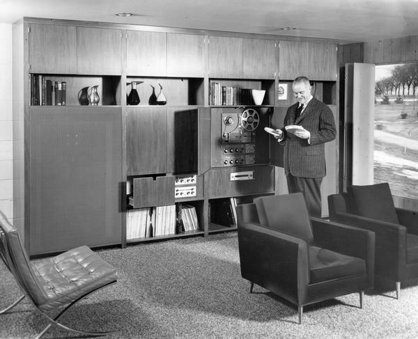 Urbane gentleman shows off his sophisticated high fidelity wall system with reel-to-reel tape deck, reels of tape, and record albums.