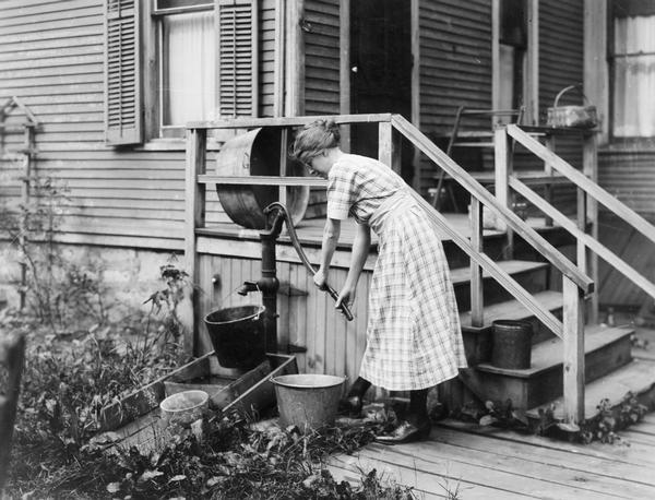 Woman pumping water from a well near the porch of her farmhouse.