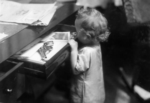 Small child opening a desk drawer containing a shining revolver (hand gun). The scene was staged for International Harvester's Agricultural Extension Department. The photograph was used to warn families of potential farm hazards. The original caption reads: "the revolver in desk drawer is in a bad place."