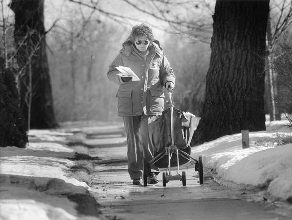 A woman mail carrier pushes her cart with a full mailbag along a sidewalk edged by snowy lawns.