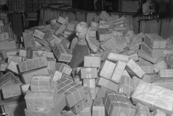Fred Boyle, clerk at the post office, looks overwhelmed surrounded by a mountain of Christmas parcels to be sorted for shipment overseas to men and women in service.