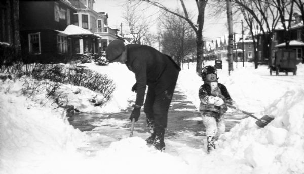 Winter scene with a man and young boy shoveling snow from a sidewalk after a heavy snowfall.