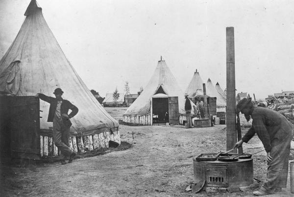 Two men cook at griddles while another leans on an open door to one of many tents at a Civil War army camp.