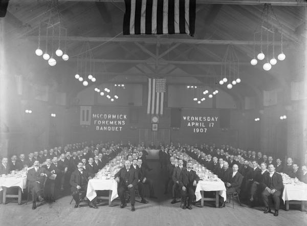 Large group of men sitting and posing at long dining tables in the Club House of International Harvester's McCormick Works factory. The men are attending the "McCormick Foreman's Banquet."