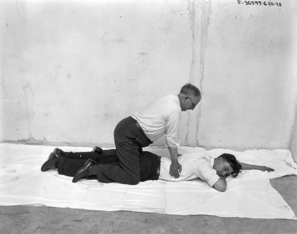 Man massaging the back of another man in a bare concrete room. The recipient of the massage is stretched out face down on the floor and the other man is kneeling over him.