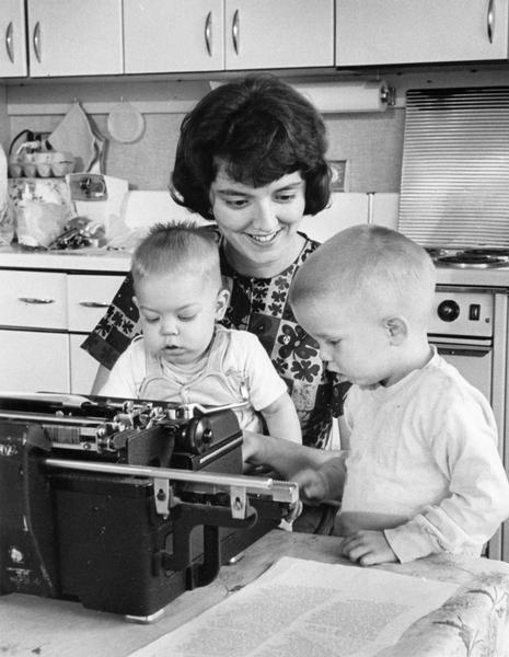 A woman and two small boys are sitting at a typewriter in the kitchen.
