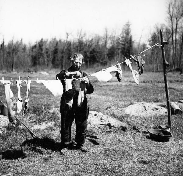 In a photograph taken for the Farm Security Administration, a man hangs socks to dry on a clothesline on land where trees have been cut down, or cutover.