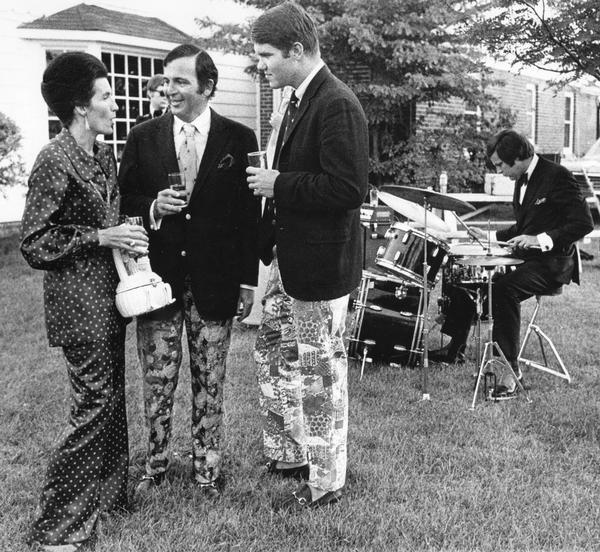A woman in a polka-dotted suit chats with two men wearing wildly-patterned slacks and sport coats at a summer party.  A man plays drums in the background.