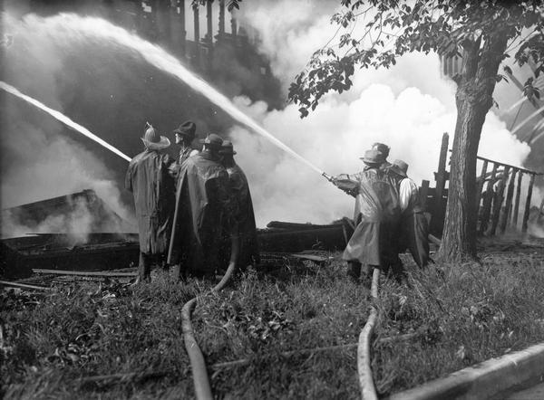 Fire fighters standing amid smoking timbers aim their hoses to douse the flames.