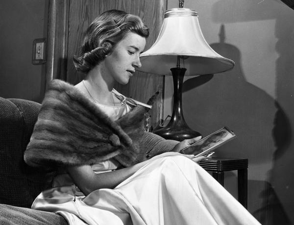 A young woman wearing a formal gown and mink stole looks at a portrait of a woman while sitting on a couch, as if waiting to go out for the evening.