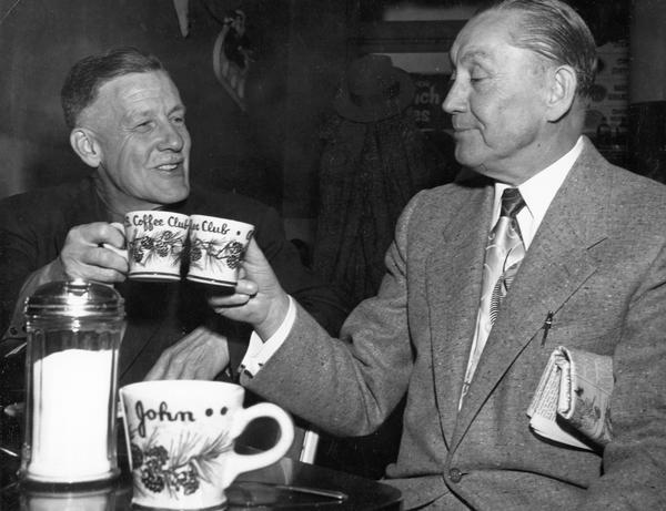 Two men clink coffee mugs as they prepare to drink their morning coffee.