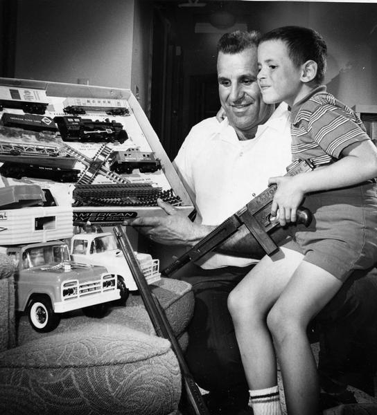 Boy holds toy gun and looks at electric train set and toy train with older man.