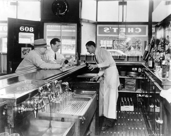 Two patrons and bartender at Chet's tavern. The bartender is pulling bottles of Schlitz beer out of an International No. 4 De Luxe dry type beverage cooler.