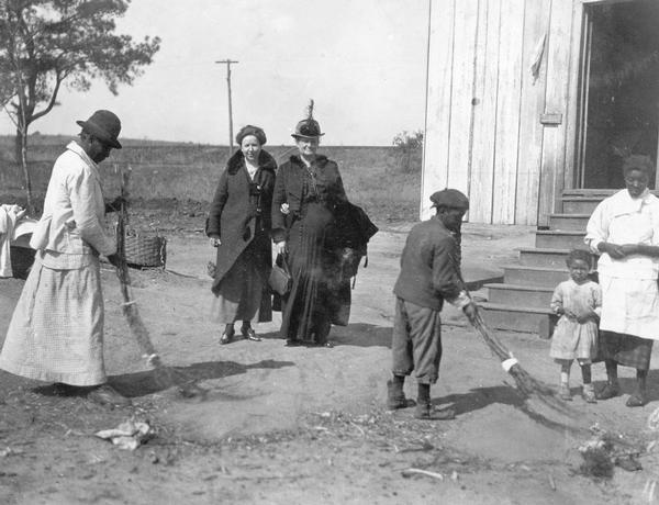 A woman and boy are using "sedge brush" brooms to sweep a dirt yard as two women — possibly social workers or Agricultural Extension employees — look on. Another woman with two children is standing near the steps to the house. The brush broom, made by tying a bundle of brush together, was used in the south for rough sweeping around the yard.