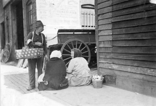 One man and two women on an urban sidewalk with baskets of produce. The man's basket is decorated with white stars on a dark field. The people are probably selling the produce on the street.