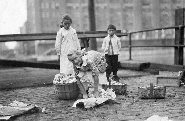 Young girl wrapping or unwrapping produce from baskets on an urban street, possibly in Chicago, Illinois. The girl is likely offering the produce. A boy and girl are standing behind her looking on.