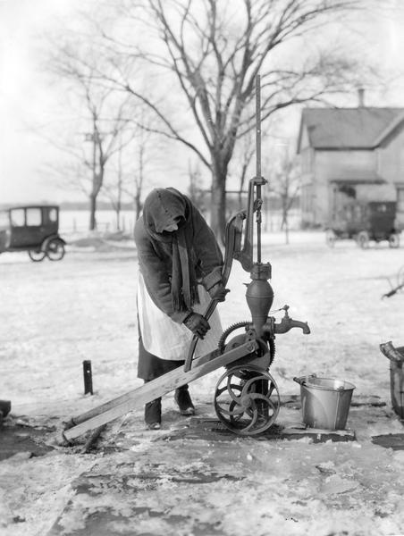 A bundled-up woman hand pumps water from a well into a pail. The ground is covered with ice and snow. Two cars and a farmhouse are in the background.