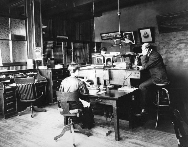 Two men and a woman working in a branch(?) office of the International Harvester Company. One man is talking on a telephone. Portraits of William Deering and Cyrus McCormick are hanging on a wall, along with the poster "Midnight at McCormick's."