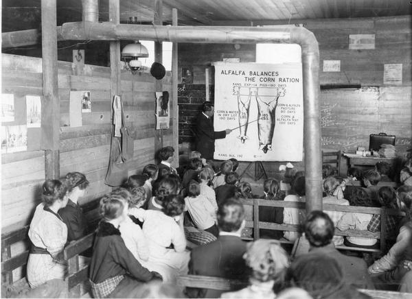 International Harvester Agricultural Extension Department employee J.E. Waggoner delivers a lecture to children and farmers assembled at Providence School. A lecture chart titled "alfalfa balances the corn ration" helps Waggoner tell the story of the corn fed pig and of his twin brother who liked alfalfa with his corn. The school house has a partition to make two rooms.