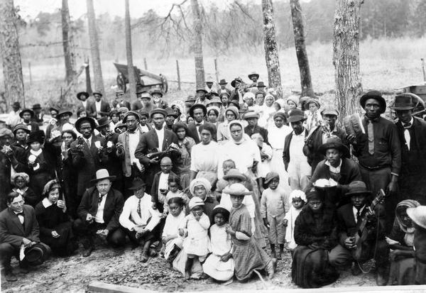African American tenant farmers assembled for a barbecue. The tenants worked for Louis Frank Sessions who is in the front row, third from the left (wearing a light-colored hat). To his right is Miss Helen Holden of Chicago. The barbecue followed a lecture by International Harvester's Agricultural Extension Department. According to the original caption, the tenants "finished up the barbecue after the white folks had eaten their fill." One man is holding a guitar and another is holding a violin (fiddle).