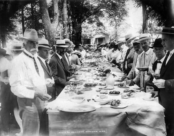 View down long table set up outdoors for a feast. Men are standing on both sides, and a young girl is standing between two men on the right.
