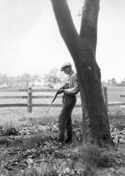 Boy idling away time with a rifle under a tree on the grounds of a rural school.