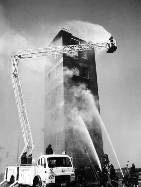 Fire fighters use an International Model VCO-202 fire and rescue truck with an eighty-five foot mounted ladder to fight a fire, possibly as part of a training exercise. The ladder's design incorporates a hose in its nose and a jointed boom. The truck was owned and operated by the Baltimore Fire Department.
