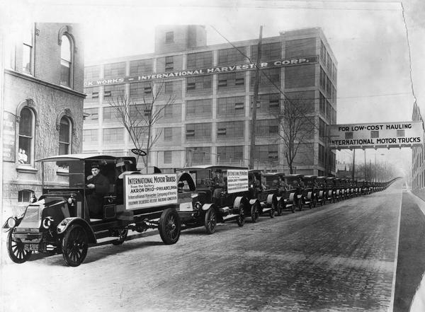 Long line of International trucks on a road next to International Harvester's Akron Works. Placards on the first two trucks read: "International motor trucks from the factory, Akron, Ohio, to Philadelphia. International Harvester Company of America; highway deliveries relieve railroad congestion." The factory was owned by the Aultman & Miller Buckeye Company until 1905, when it was purchased by International Harvester.