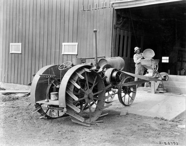 Man standing inside the doorway of a barn pouring feed into a feed grinder. The grinder is powered by a belt attached to a Titan 10-20 tractor.