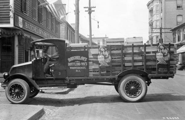 International Model G-61 loaded with Orange Smash soda and parked in middle of a city street. A man is sitting in the driver's seat. The truck was owned and operated by Batchelor's Bottling Works.