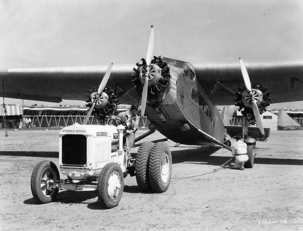 Crew preparing to pull a damaged Ford Tri-Motor airplane with a white McCormick-Deering I-30 industrial tractor with dual rear wheels at the American Air Races. The races took place at Chicago's Municipal Airport, July 1-5, 1933. A man hitching a tow line to the plane is wearing a jumpsuit with the words "Aeronautical University" on the back.