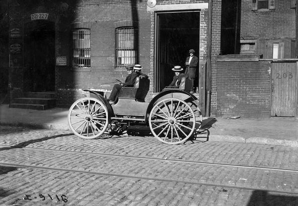 Three men sit in an International Auto Buggy parked on an urban cobblestone street while a fourth man looks on from the doorway of a brick building. A small sign on a nearby doorway reads: "Westinghouse Electric & Manuf'g Company."