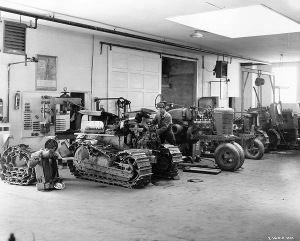 Mechanic taking apart a tractor engine in the service department of the Snake River Equipment Company, an International Harvester dealership. An International diesel TD-6 TracTracTor (crawler tractor), a Farmall M and three other unidentified industrial and farm tractors are also in the shop. The store was managed by George Watkins.