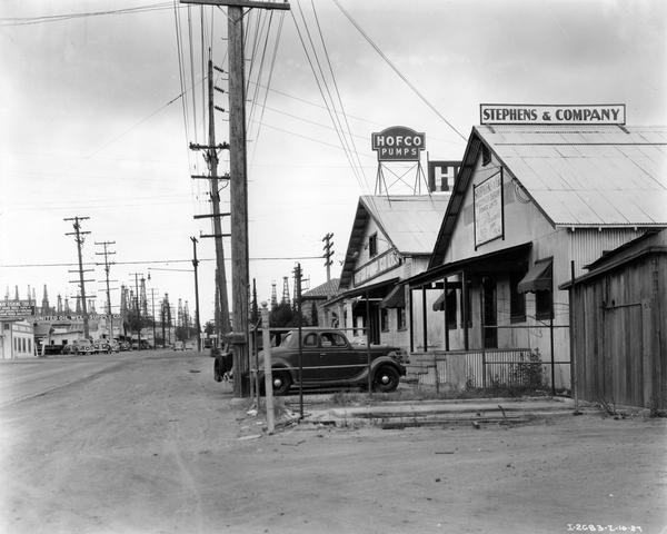 Businesses and cars parked along a dusty city street with oil derricks in the background. At right is the Stephens & Company, distributors of power units in California oil fields, and the Hofco Pump Co.