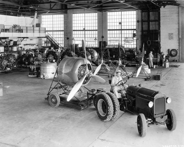 Slightly elevated view of a worker moving an airplane engine with an International I-30 industrial tractor and trailer inside the Municipal Airport hangar. The tractor was owned by American Airlines.