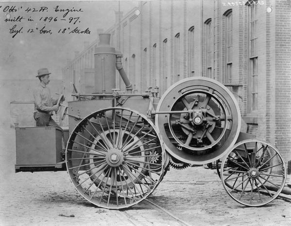 Side view of a man posing standing and holding onto the steering wheel of an "Otto 42 HP engine, which was built in 1896-97" with a 12-inch bore and 18-inch stroke.
