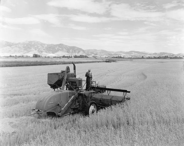 Elevated view of men harvesting grain with a McCormick-Deering 12 foot experimental combine (harvester-thresher) pulled by a Farmall M tractor. There is a mountain range in the background.